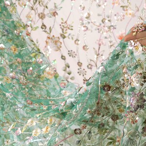 a close-up view of a hand holding a piece of fabric with a floral pattern. The fabric appears to be a light green material with a variety of colorful flowers and leaves, creating a visually appealing and intricate design.