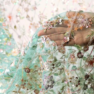 A hand is holding a piece of fabric with intricate floral embroidery, including various shades of green, pink, and white flowers and leaves.