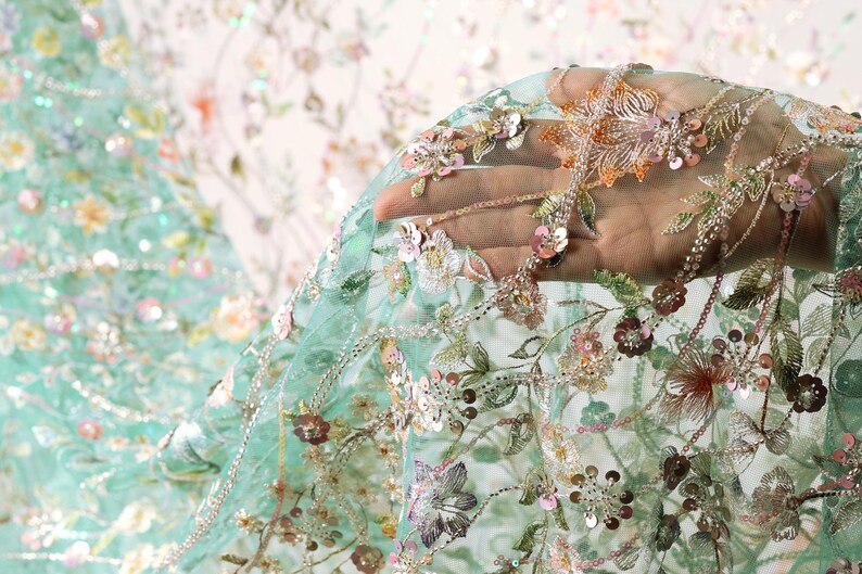 A hand is holding a piece of fabric with intricate floral embroidery, including various shades of green, pink, and white flowers and leaves.