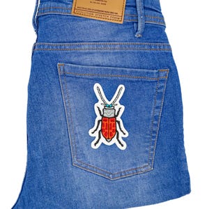Peut inclure: Un jean bleu denim avec un patch en cuir marron qui dit "PULL&BEAR EVOLUTION STRETCH DENIM" et un patch brodé d'un insecte rouge et blanc sur la poche arrière.