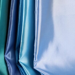 May include: A collection of folded satin fabric sheets arranged in a vertical gradient. The colors transition from a deep teal on the left to a vibrant royal blue, a bright sky blue, and finally a pale, light blue on the far right side.