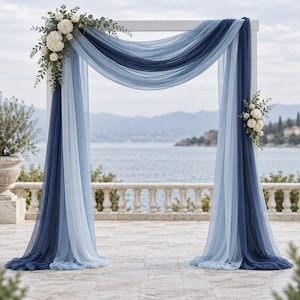 May include: A white rectangular wedding arch draped with layers of sheer fabric in shades of navy blue and light blue. The structure is decorated with clusters of white flowers and green eucalyptus leaves on the top left and middle right corners.