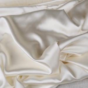 Könnte beinhalten: Ein Stück cremeweißer Stoff mit einer glatten, glänzenden und reflektierenden Oberfläche. Das Material ist in weichen, fließenden Falten drapiert, wodurch tiefe Schatten und helle Glanzlichter entstehen, die die seidige Textur des Stoffes betonen.