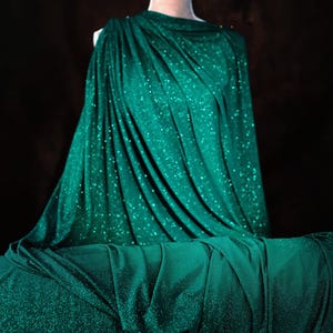 A mannequin wearing a shimmering green dress with a draped, flowing silhouette against a dark background.