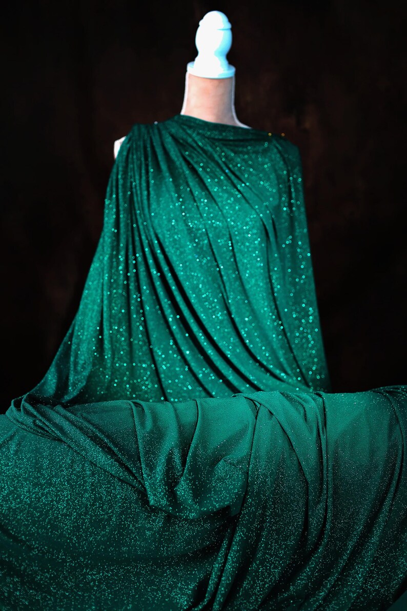 A mannequin wearing a shimmering green dress with a draped, flowing silhouette against a dark background.