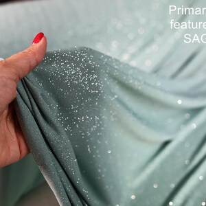 A hand holding a piece of fabric with a glittery texture, and the background is a blurred green color.