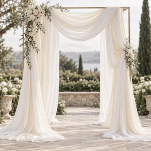 May include: A rectangular gold metal frame arch draped with flowing white sheer fabric. The structure is decorated with clusters of white roses and green foliage attached to the top left corner and the middle right side of the metal frame.