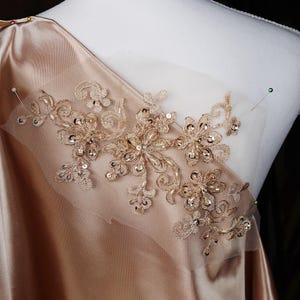 Może przedstawiać: Złota aplikacja z koronki z cekinami, przypięta do jasnobrązowej satynowej tkaniny. Aplikacja przedstawia kwiatowy wzór z delikatnymi detalami.