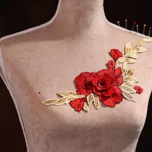 Puede incluir: Una aplicación floral roja y dorada con una rosa grande en el centro. La aplicación está fijada a un maniquí cubierto de tela.