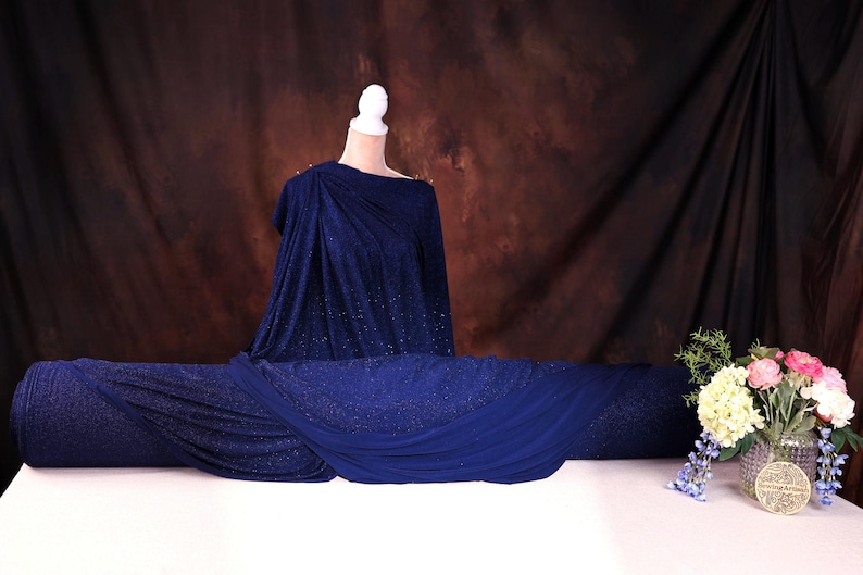 a mannequin wearing a blue dress with a draped, flowing design, placed on a white table. Next to the mannequin is a vase filled with flowers, creating a visually appealing and elegant display.