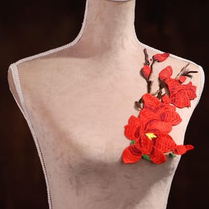 Puede incluir: Aplique floral bordado en rojo con hojas verdes y tallos marrones. El aplique está sobre un fondo de tela marrón claro.