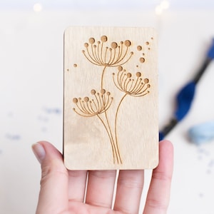 Könnte beinhalten: Eine Holzkarte mit einem Laser-geschnittenen Design von drei stilisierten Löwenzahnpflanzen mit runden Samenköpfen.