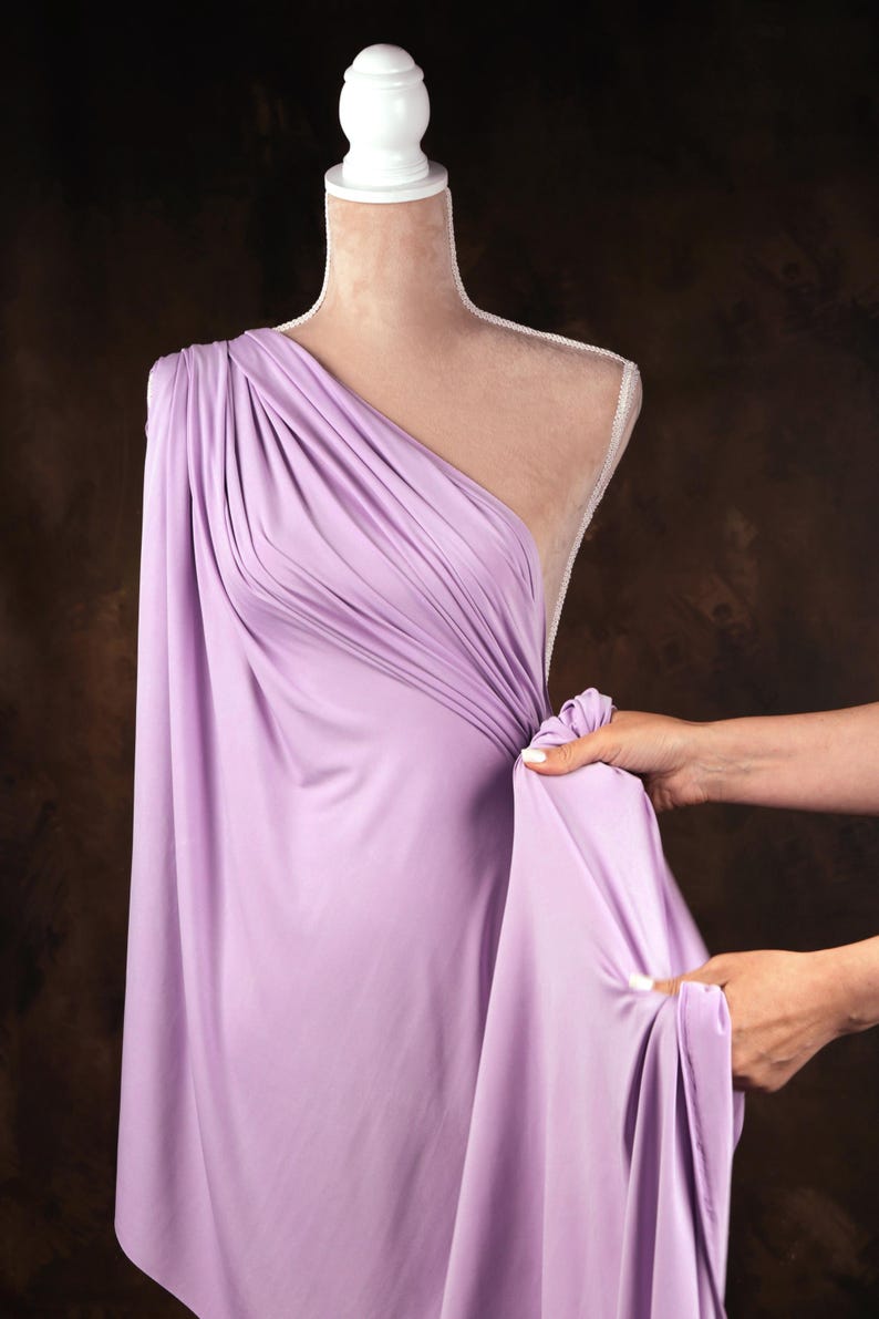 A mannequin wearing a lavender one-shoulder dress is being adjusted by someone's hands.
