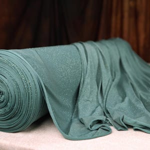 A roll of green fabric with a sparkly texture is placed on a table, with a vase of pink flowers in the background.