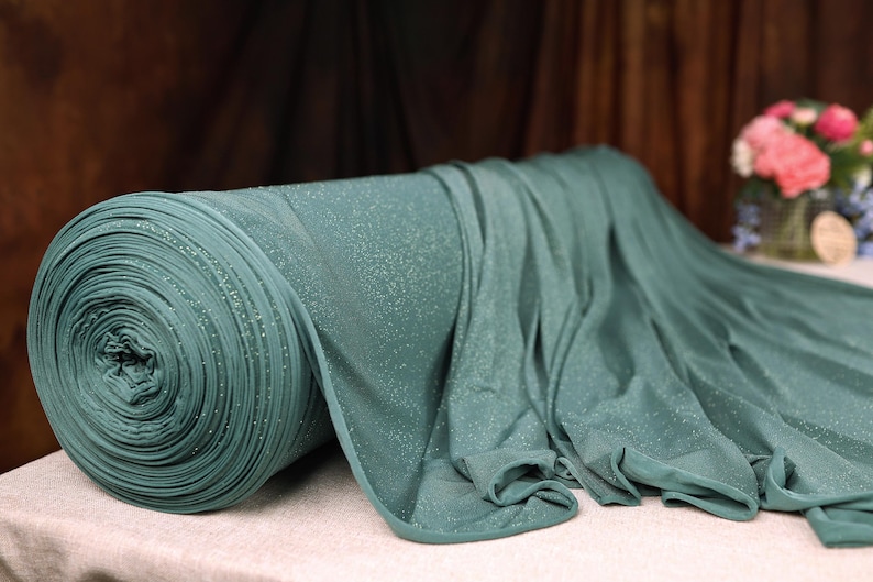 A roll of green fabric with a sparkly texture is placed on a table, with a vase of pink flowers in the background.