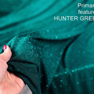 A hand holding a piece of fabric with a shimmering green color, likely Hunter Green, against a blurred teal background.