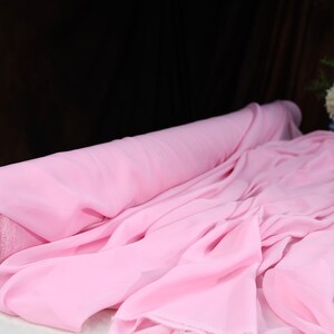 A roll of pink chiffon fabric is displayed on a table, with a vase of flowers in the background.