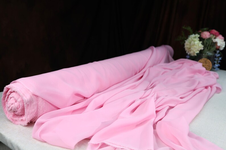 A roll of pink chiffon fabric is displayed on a table, with a vase of flowers in the background.