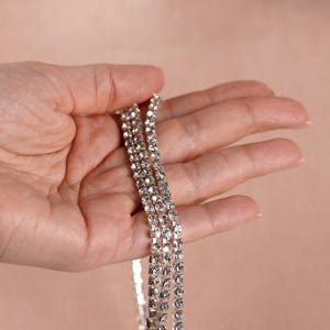 May include: A hand holding three strands of a silver rhinestone necklace. The necklace is made of small, sparkling rhinestones set in a silver metal. The background is a soft, neutral color.