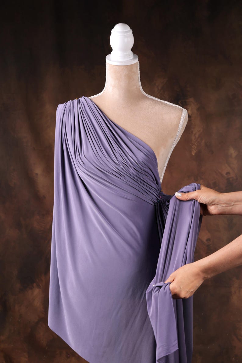 A mannequin wearing a purple one-shoulder dress is being adjusted by someone's hands.