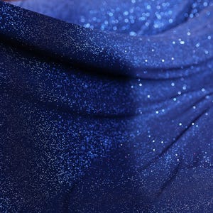 The image is a close-up of a shiny, glittery fabric with a blue color. The fabric appears to be made of a shiny material, possibly sequin or glittery fabric, and has a sparkling texture. The background of the image is dark blue, which contrasts with the blue color of the fabric and adds depth to the composition.