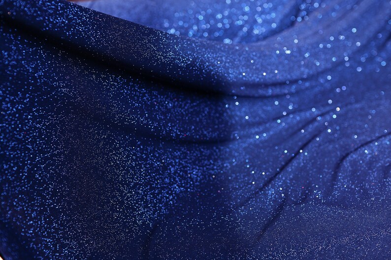 The image is a close-up of a shiny, glittery fabric with a blue color. The fabric appears to be made of a shiny material, possibly sequin or glittery fabric, and has a sparkling texture. The background of the image is dark blue, which contrasts with the blue color of the fabric and adds depth to the composition.