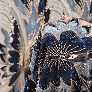 Puede incluir: Primer plano de una tela azul marino y azul claro con adornos dorados y negros. La tela presenta un diseño floral con lentejuelas y abalorios. El fondo muestra un patrón similar en una paleta de colores más clara.