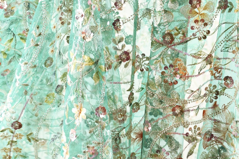 The image is a close-up of a fabric with a floral pattern, featuring various shades of green, pink, and yellow flowers and leaves. The fabric appears to be made of a sheer material, giving it a delicate and airy appearance.