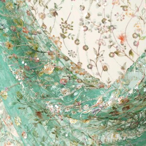 a close-up view of a fabric with a floral pattern, including various shades of green, pink, and white flowers and leaves. The fabric appears to be made of a sheer material, giving it a delicate and airy appearance.