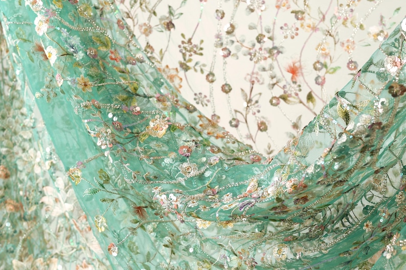 a close-up view of a fabric with a floral pattern, including various shades of green, pink, and white flowers and leaves. The fabric appears to be made of a sheer material, giving it a delicate and airy appearance.