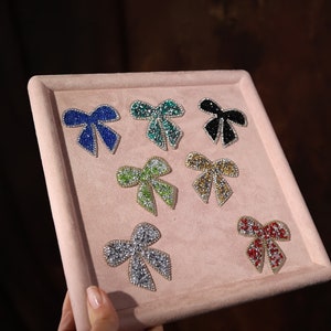 Puede incluir: Una muestra de nueve lazos de pedrería en varios colores, incluyendo azul, verde, dorado, plateado y rojo, sobre un fondo de terciopelo rosa.