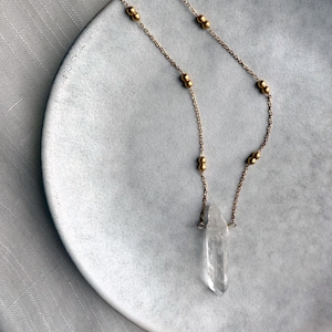 May include: A delicate gold-toned necklace featuring a clear quartz crystal pendant. The necklace has a fine chain interspersed with small, round beads. The crystal is elongated and pointed, adding a natural, elegant touch to the jewelry.