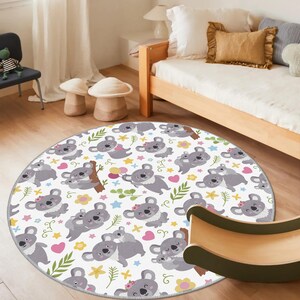 May include: A round, white rug featuring a repeating pattern of gray koalas with pink flowers, hearts, and colorful stars. The rug has a gray border and is designed for a child's room.
