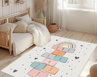 May include: A rectangular area rug featuring a hopscotch game design with numbered squares from one to nine. The rug has a white background with small gray dots and dark gray hearts, and a rainbow graphic at the top above the number nine.