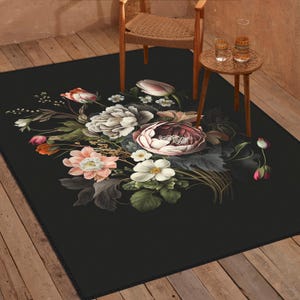 May include: A rectangular area rug featuring a black background with a central floral arrangement of white, pink, and peach-colored flowers and dark green leaves. The rug is placed on a wooden floor beneath a wooden chair and a small round side table.