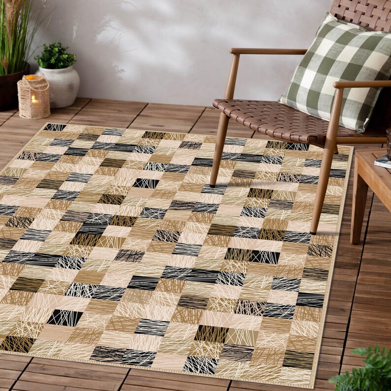 May include: Rectangular area rug with a geometric pattern in shades of beige, tan, and black. The rug features a patchwork design with textured lines and shapes, creating a modern and stylish look. The rug is placed on a wooden deck.