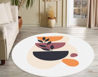 May include: A round area rug featuring a white background with a central graphic design. The design includes three stacked semi-circles in orange, maroon, and dark blue, with a black leafy branch overlay and additional abstract orange and maroon shapes.