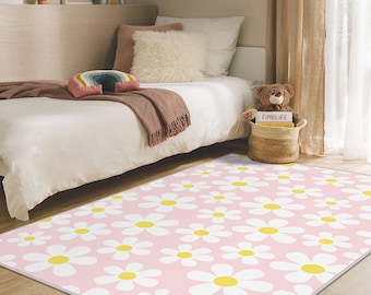 May include: A pink rectangular rug featuring a daisy pattern in white and yellow. The rug is in a bedroom setting, with a bed, pillows, and a teddy bear in the background. The rug's dimensions are approximately 152 cm by 213 cm.