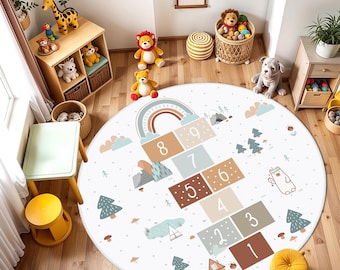 May include: A round area rug featuring a hopscotch game pattern with numbers one through nine in muted earth tones. The design includes illustrations of mountains, pine trees, a small house, and a bear, all set against a white background with dots.