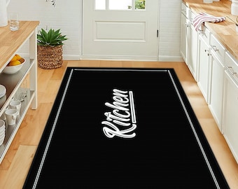 May include: A rectangular black floor mat featuring a white border and the word Kitchen written in a stylized white script font in the center. The mat has a clean, simple design with a solid black background and white text and border accents.