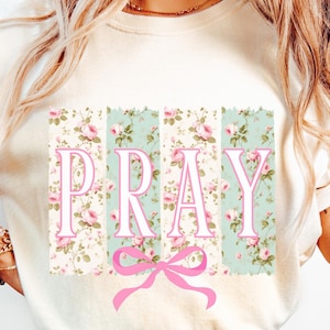 May include: A white t-shirt with a floral print and the word "PRAY" in pink letters. A pink bow is centered below the word.