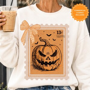 Pode incluir: Uma camisola branca de manga comprida com um gráfico de selo postal que mostra uma abóbora esculpida, pequenos morcegos e estrelas. O design inclui um laço de xadrez laranja claro no canto superior esquerdo e o texto 13c no canto superior direito.