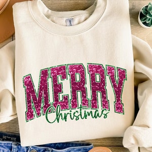 以下が含まれることがあります： キラキラ光るピンクとグリーンのデザインで「MERRY Christmas」と書かれたクリーム色のスウェットシャツ。スウェットシャツは木製の表面に折りたたまれており、背景には帽子とジーンズがあります。小さな多肉植物も見えます。