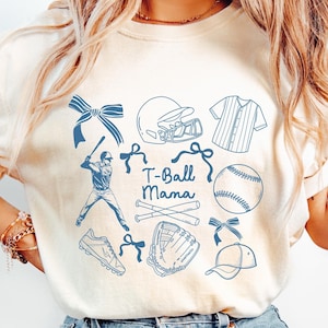 May include: Cream-colored t-shirt with a blue baseball-themed graphic. The design includes a baseball player, baseball equipment, and the text "T-Ball Mama". The shirt has short sleeves and a crew neck.