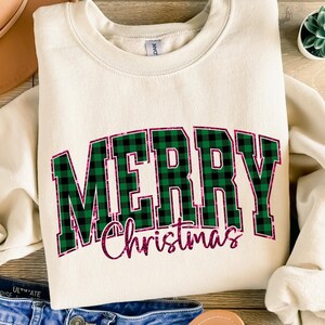 May include: Cream-colored sweatshirt with the words "MERRY Christmas" in a green and black plaid design with a pink outline. The sweatshirt is folded, with a brown hat and blue jeans in the background.