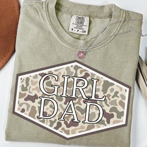 May include: An olive green t-shirt with the words "GIRL DAD" in a white font inside a brown and tan camouflage hexagon. The shirt has a Comfort Colors tag and a silver necklace with a pink pendant.