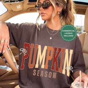 Pode incluir: Uma t-shirt de manga curta castanho-escura com o texto PUMPKIN SEASON em letras grandes. A palavra PUMPKIN está decorada com bolinhas e pequenos laços brancos. Um emblema circular verde indica COMMERCIAL LICENCE INCLUDED.