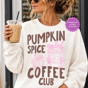 May include: A white long-sleeve crewneck sweatshirt featuring the brown text PUMPKIN SPICE COFFEE CLUB and a pink graphic illustration of a coffee cup and a pumpkin surrounded by leaves. The sweatshirt is worn with blue denim jeans.