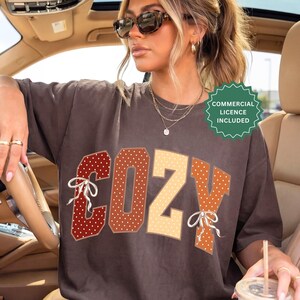 Pode incluir: Uma t-shirt de manga curta castanho-escura com a palavra COZY em letras grandes de estilo bloco com um padrão de bolinhas brancas. As letras são coloridas em tons de vermelho, laranja e creme, com pequenos laços de fita branca no C e Y.