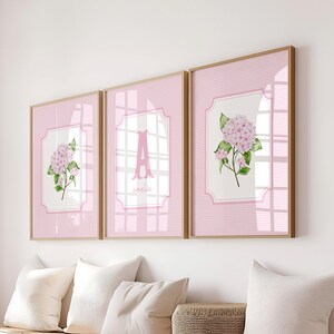 May include: Three framed wall art pieces with a pink gingham pattern background. Each frame has a light pink hydrangea flower illustration. The center frame features the letter "A". The frames are displayed above a bench with white pillows.